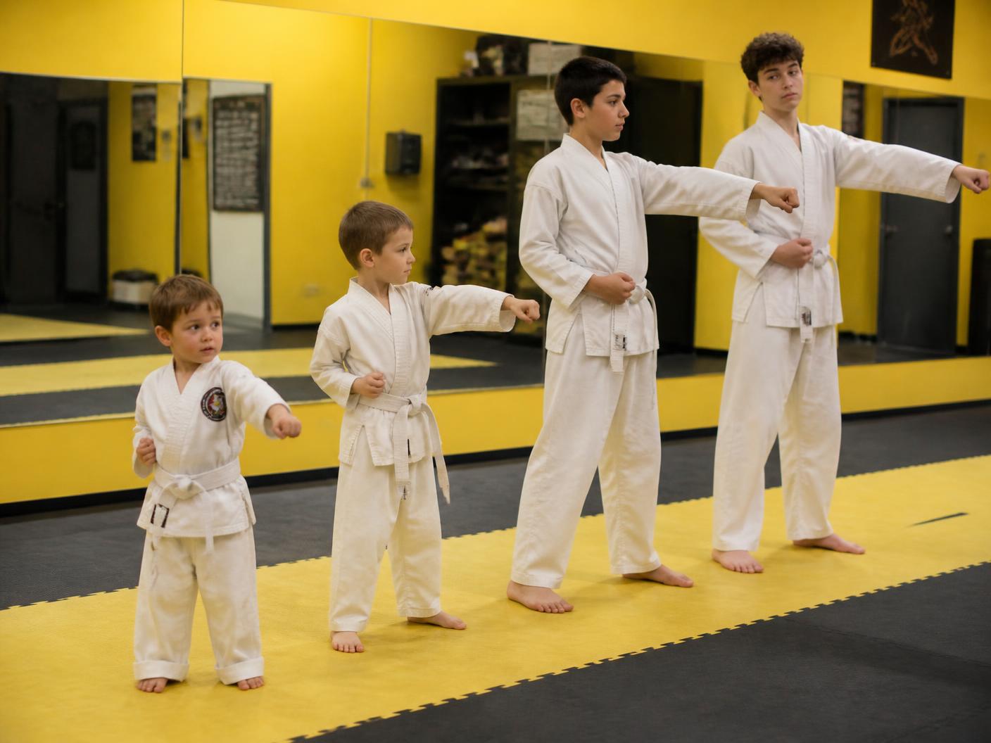 A preschooler and a pre-teen standing side by side in the same martial arts line, being taught the same drill